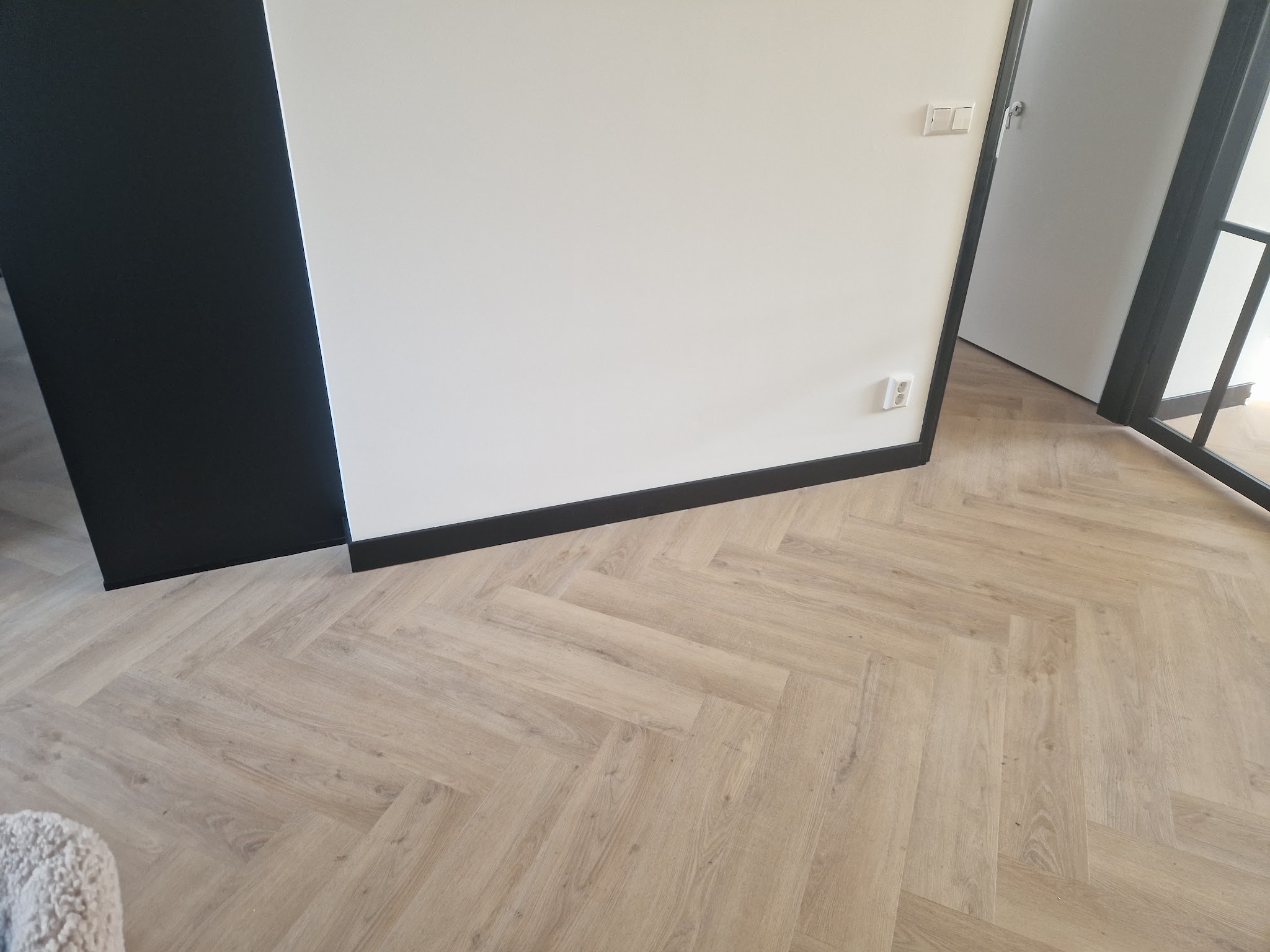 Lichte visgraatvloer langs een witte wand met zwarte plint